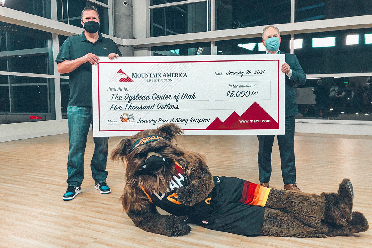 two men holding a donation check with the Utah Jazz mascot laying on the ground in front